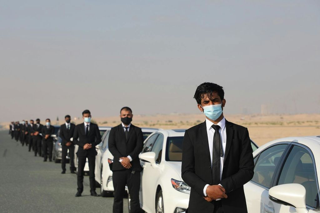 사막 풍경의 포장 도로에 가면을 쓴 남성들이 주차된 흰색 자동차 옆에 줄지어 서 있습니다.