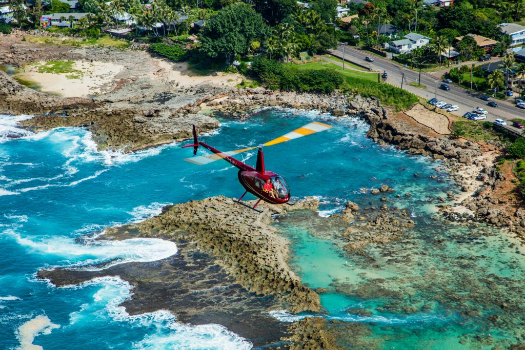 붉은 헬리콥터가 바위투성이의 해안선과 청록색 바다, 도로와 푸른 나무 위를 날고 있습니다.