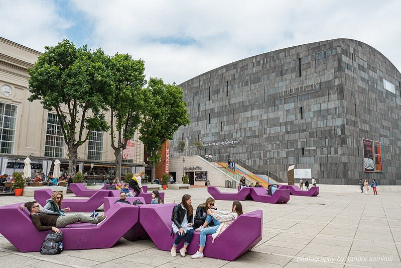 커다랗고 회색빛 건물과 나무를 배경으로 한 탁 트인 광장의 커다랗고 보라색의 현대적인 벤치에 앉아 있는 사람들.