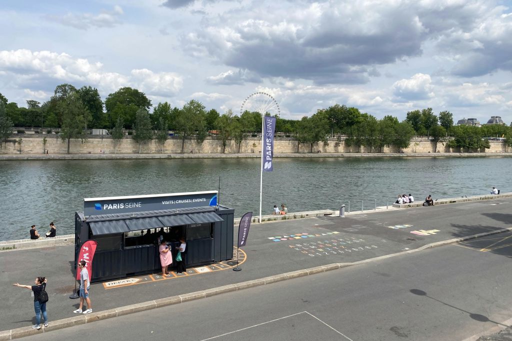 "PARIS SEINE" 브랜딩이 있는 강변 부스, 인근의 사람들, 부분적으로 흐린 하늘 아래 나무를 배경으로 한 강.