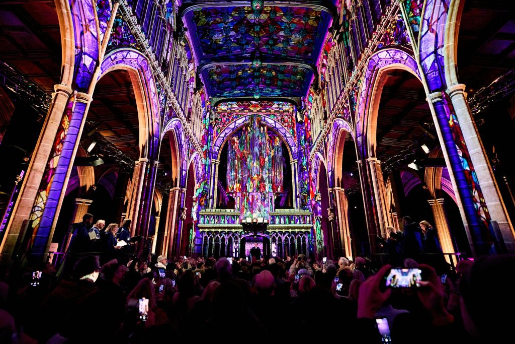 스테인드글라스 창문, 조명이 켜진 아치, 청중이 모인 활기찬 대성당 내부.