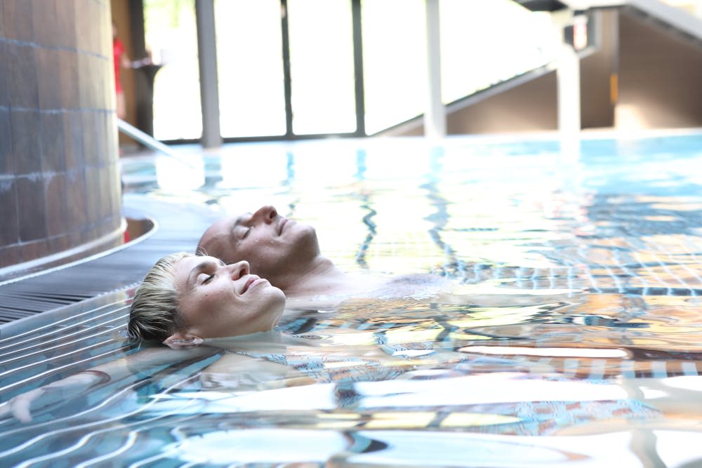 커다란 창문을 배경으로 두 사람이 눈을 감은 채 실내 수영장에서 등을 돌리고 있습니다.