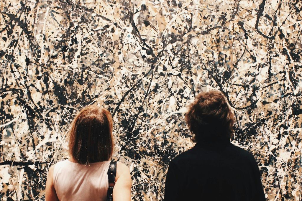 카메라를 등지고 있는 두 사람이 검은색, 베이지색, 흰색 톤의 대형 추상 튐 그림을 관찰하고 있습니다.