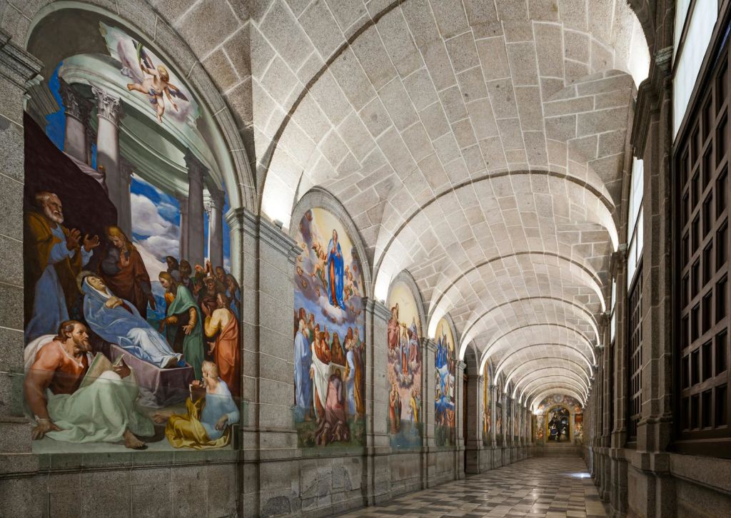 벽에 아치형 천장과 크고 화려한 종교 프레스코화가 있는 긴 석조 복도로, 다양한 성경 장면을 묘사합니다.