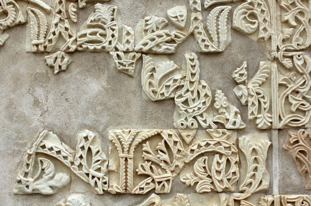 나뭇잎과 기하학적 무늬가 있는 정교하고 부분적으로 부서진 조각이 특징인 돌 벽의 클로즈업입니다.