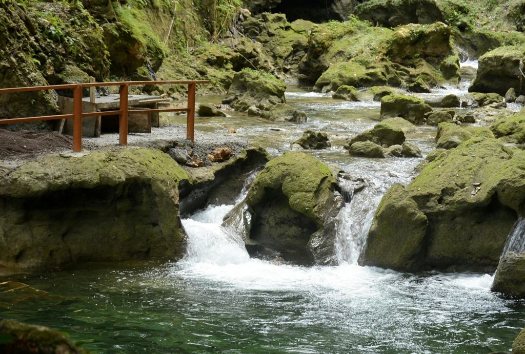 왼쪽에 나무 플랫폼 다리가 있는 이끼가 무성한 바위 위로 흐르는 물줄기. 빽빽한 녹지와 바위가 이 지역을 둘러싸고 있습니다.
