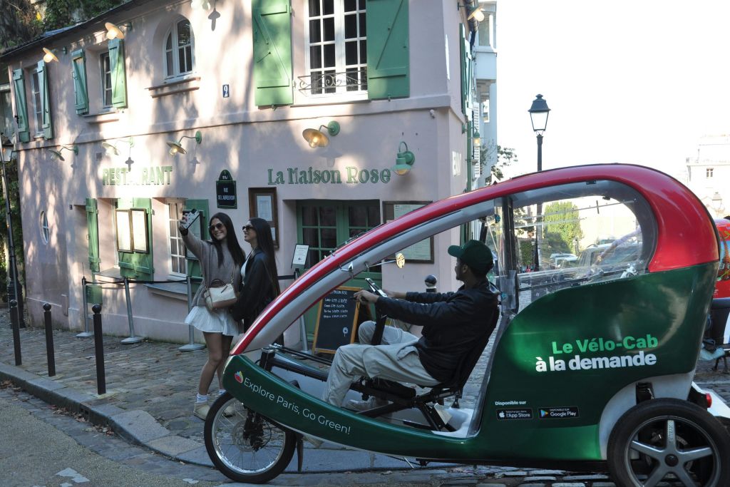 초록색 셔터가 달린 분홍색 레스토랑인 라 메종 로즈 (La Maison Rose) 외부에서 두 여성이 셀카를 찍고 있습니다. 초록색 페디캡을 탄 한 남성이 지나갑니다.