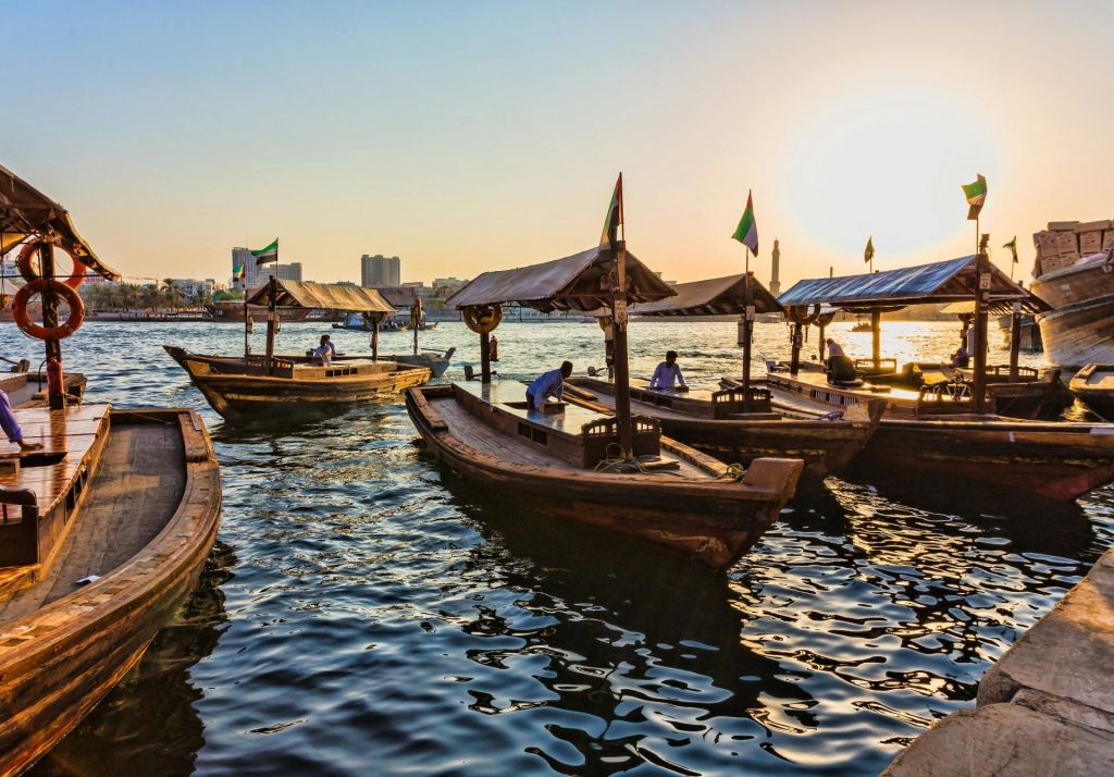 도시 건물과 모스크를 배경으로 햇살이 내리쬐는 강에 캐노피가 깔린 나무 보트가 떠 있습니다.