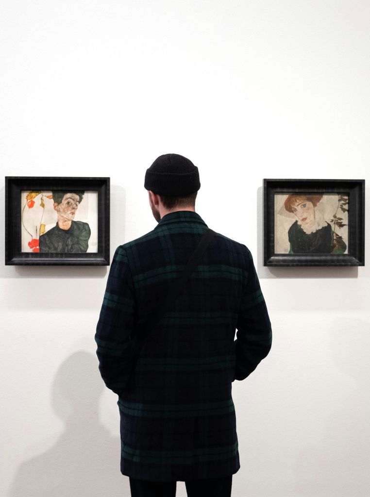 검은색 비니와 짙은 격자무늬 재킷을 입은 사람이 흰색 벽에 걸린 두 개의 액자 예술품 앞에 서 있습니다.