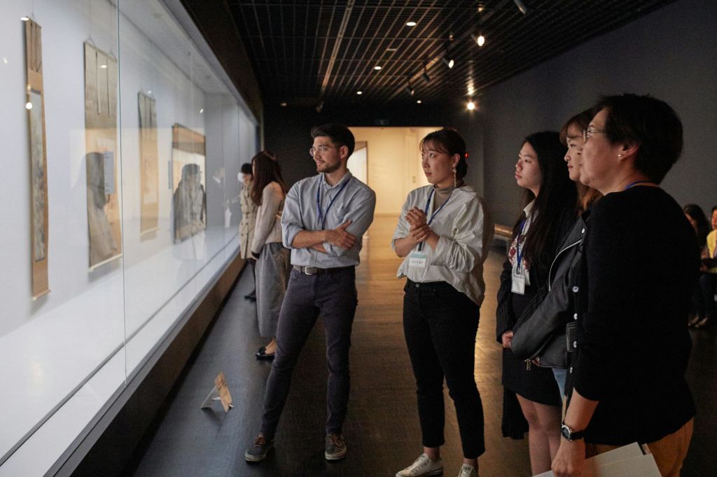 유리벽에 전시된 조명이 희미한 박물관 갤러리에서 유물을 관찰하는 사람들.