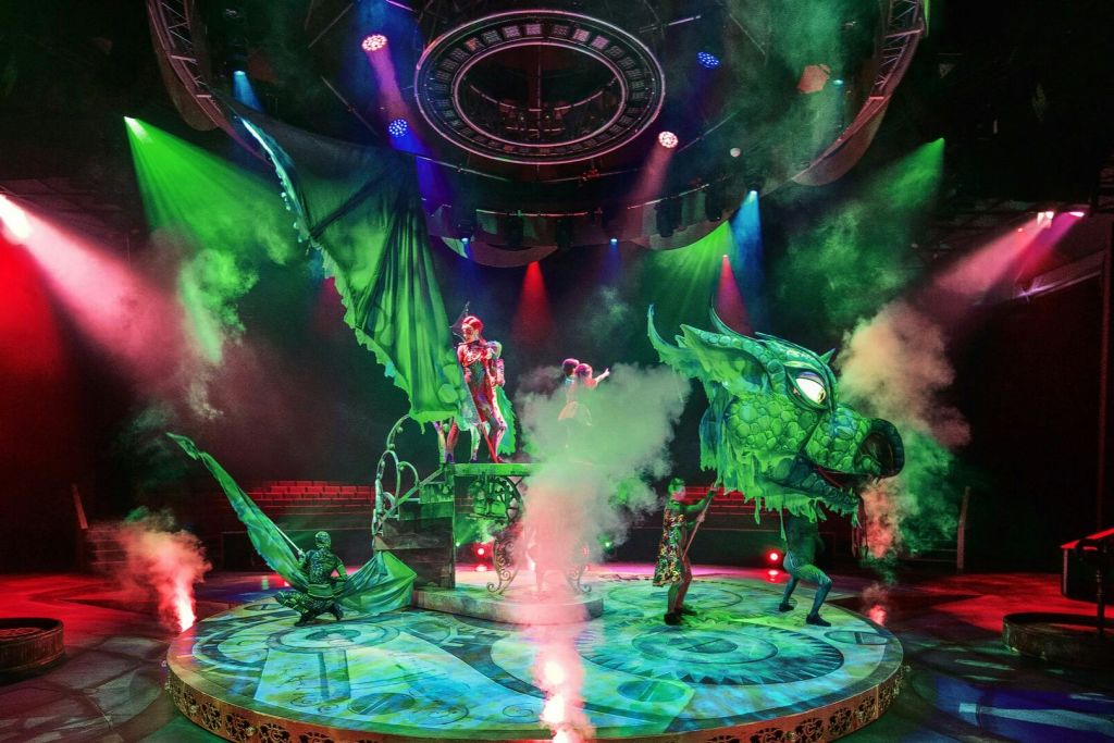 연기 효과와 화려한 조명이 있는 조명 무대에서 화려한 의상과 커다란 녹색 용 꼭두각시를 입은 공연자들.