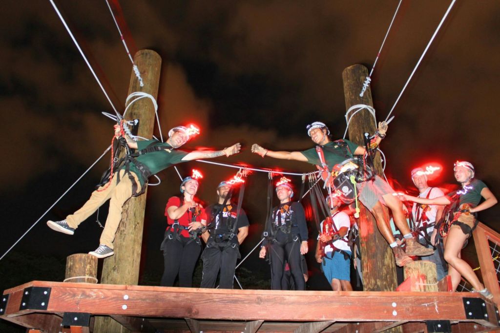 지퍼 라이닝 코스의 나무 플랫폼에 안전 장치와 헬멧에 빨간 불이 달린 사람들이 모여 있습니다.