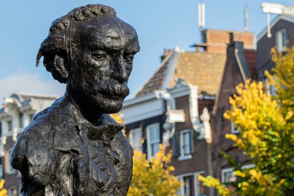 유럽 전통 건물과 노란 잎이 있는 나무를 배경으로 콧수염을 기른 남자의 청동 흉상.