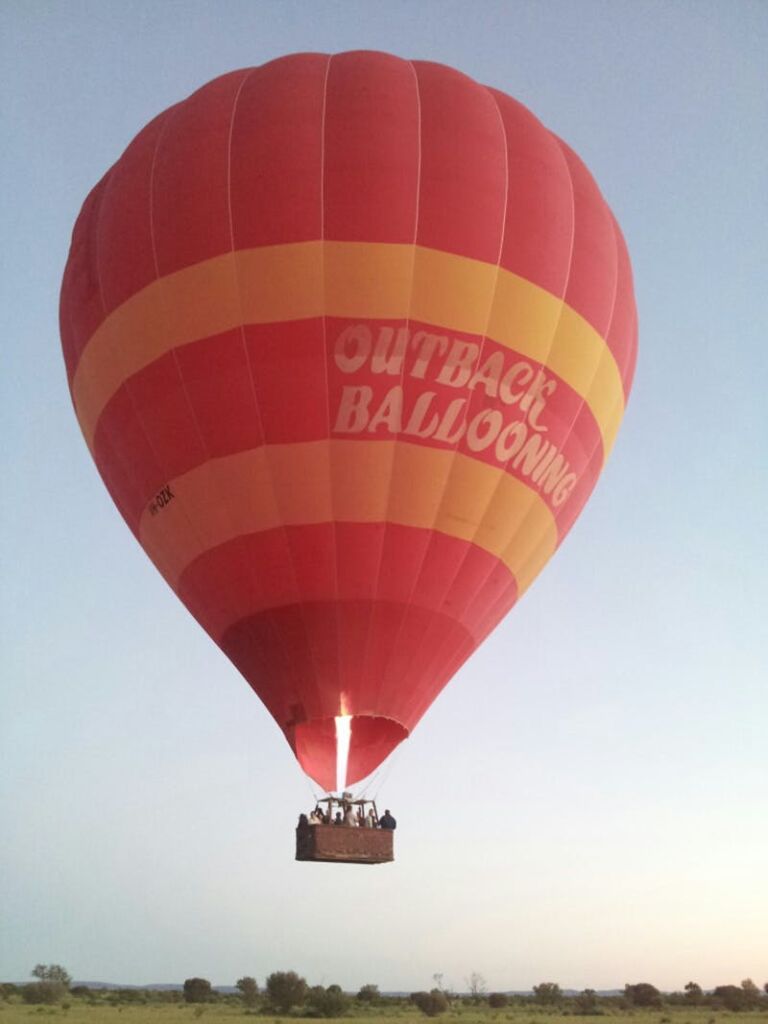 "Outback Ballooning" 텍스트가 있는 빨간색과 노란색 열기구가 사람을 태우고 맑은 하늘을 배경으로 솟아 있습니다.