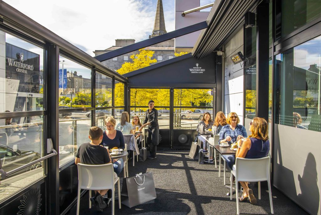 나무와 건물이 보이는 유리 벽으로 둘러싸인 화창한 날씨에 야외 테라스에서 식사를 하는 사람들.