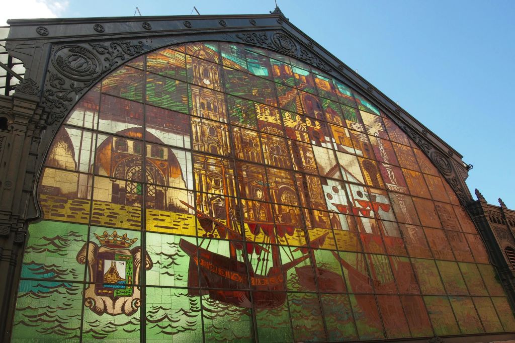 맑고 푸른 하늘 아래 화려한 구조로 장식된 역사적인 건물, 선박, 바다 요소가 묘사된 생동감 넘치는 스테인드글라스 벽화.