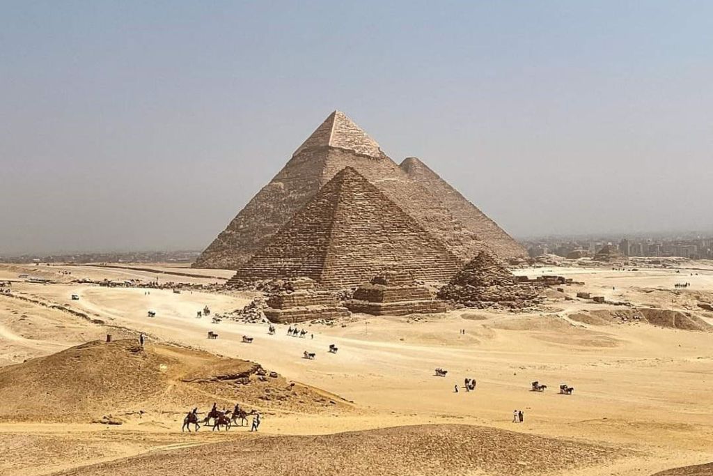 사막의 피라미드 세 개와 낙타를 타는 사람들, 그리고 맑은 하늘 아래서 근처를 걷는 사람들.