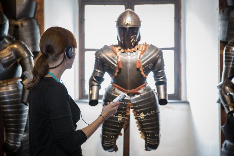 커다란 창문 앞에 진열된 갑옷을 바라보는 헤드폰과 오디오 가이드를 든 여성.