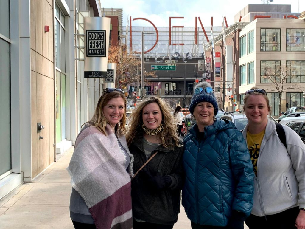 상점과 '덴버' 를 배경으로 한 커다란 간판이 있는 도시 거리에 네 사람이 미소를 짓고 서 있습니다.