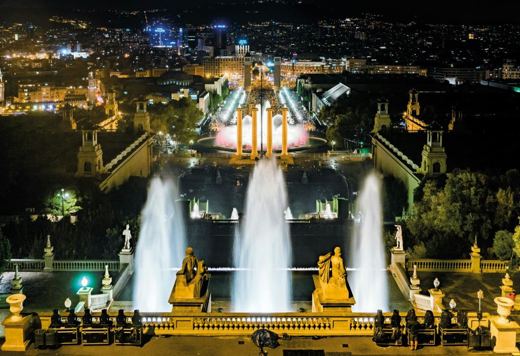 조명이 켜진 분수, 전경의 조각상, 배경에 조명이 켜진 건물이 있는 도시의 야경.