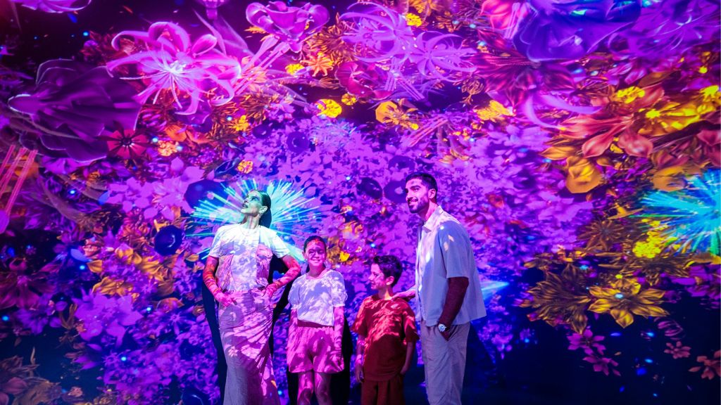 벽과 천장에 활기차고 화려한 꽃과 추상적인 디지털 투영으로 둘러싸인 4명이 함께 서 있습니다.