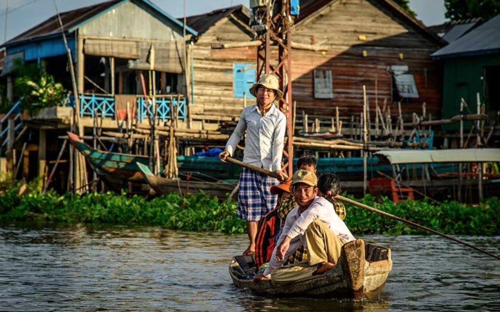 수로에 있는 나무 보트에 세 사람이 타고 있으며, 철조망을 배경으로 한 주택이 있습니다. 서 있는 남자가 노를 들고 있다.