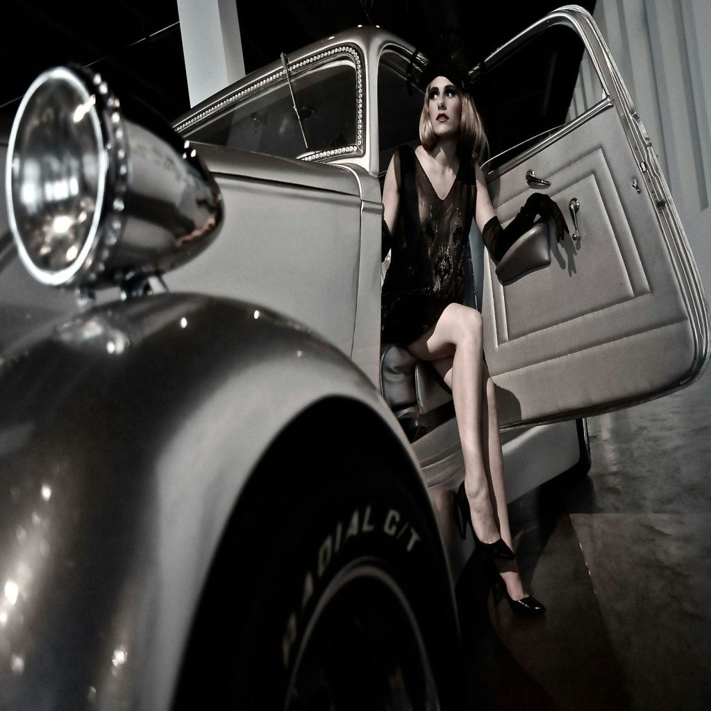 검은색 드레스와 장갑을 착용한 여성이 오픈 빈티지 차량에 앉아 전경에 헤드라이트와 타이어가 달린 채 밖으로 나가는 것처럼 보입니다.