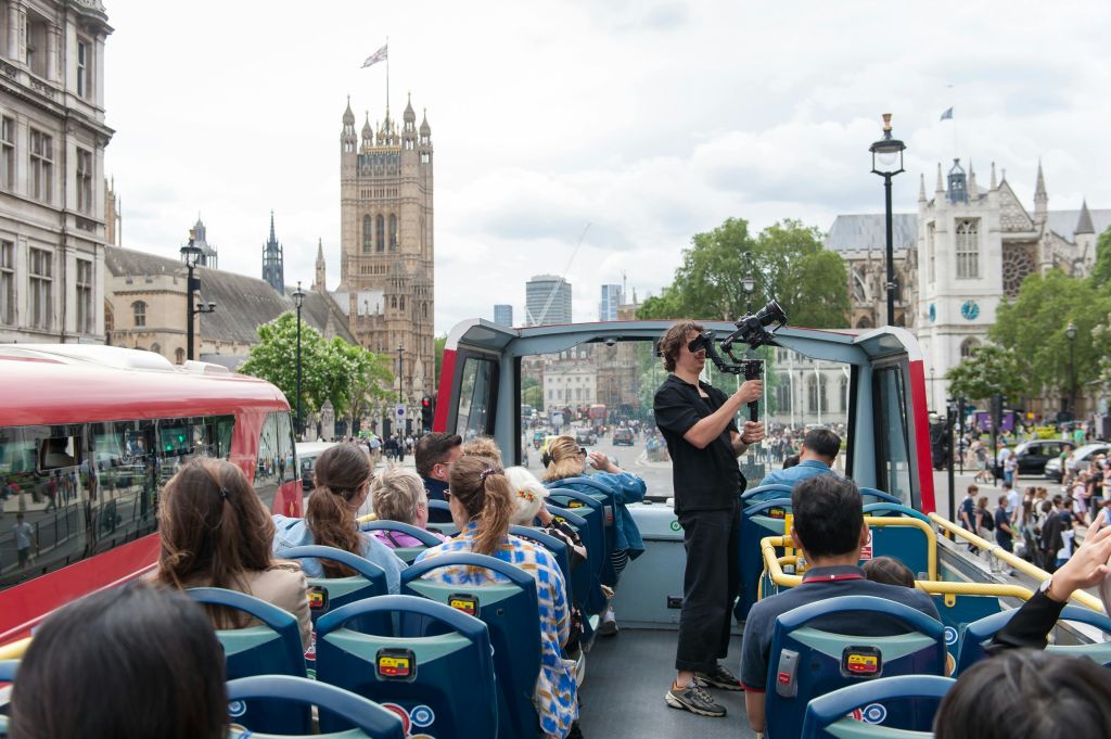 한 사람이 카메라를 이용해 촬영하는 오픈탑 버스 투어에 참여한 관광객들. 거대한 역사적 건물이 배경에 있습니다.