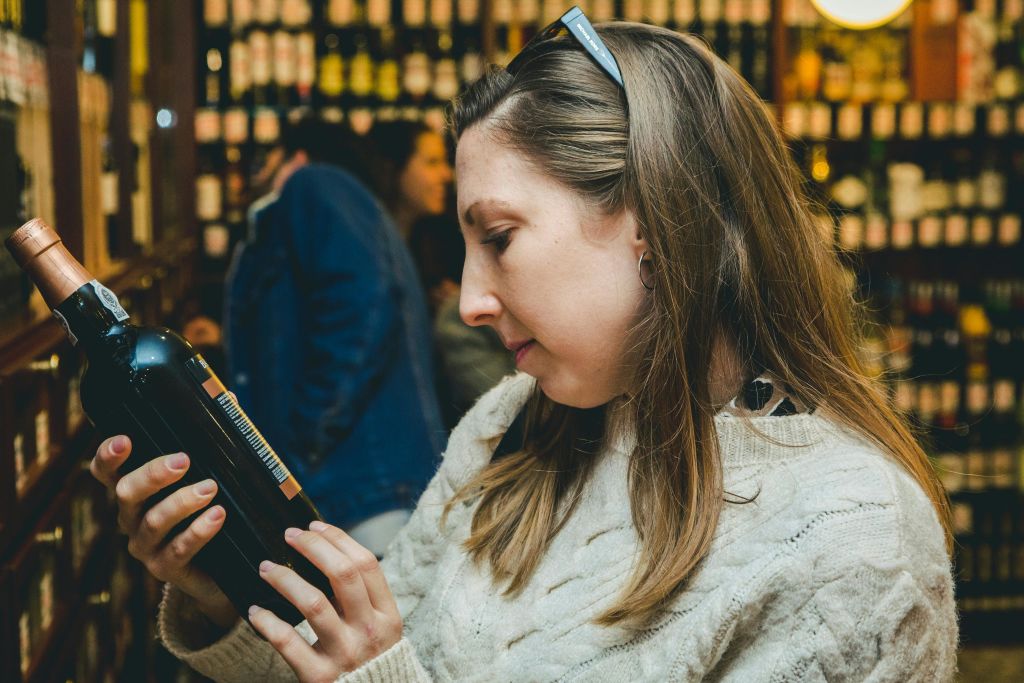 흰색 스웨터를 입은 여성이 병으로 채워진 선반 배경이 있는 매장에서 병을 살펴보고 있습니다.