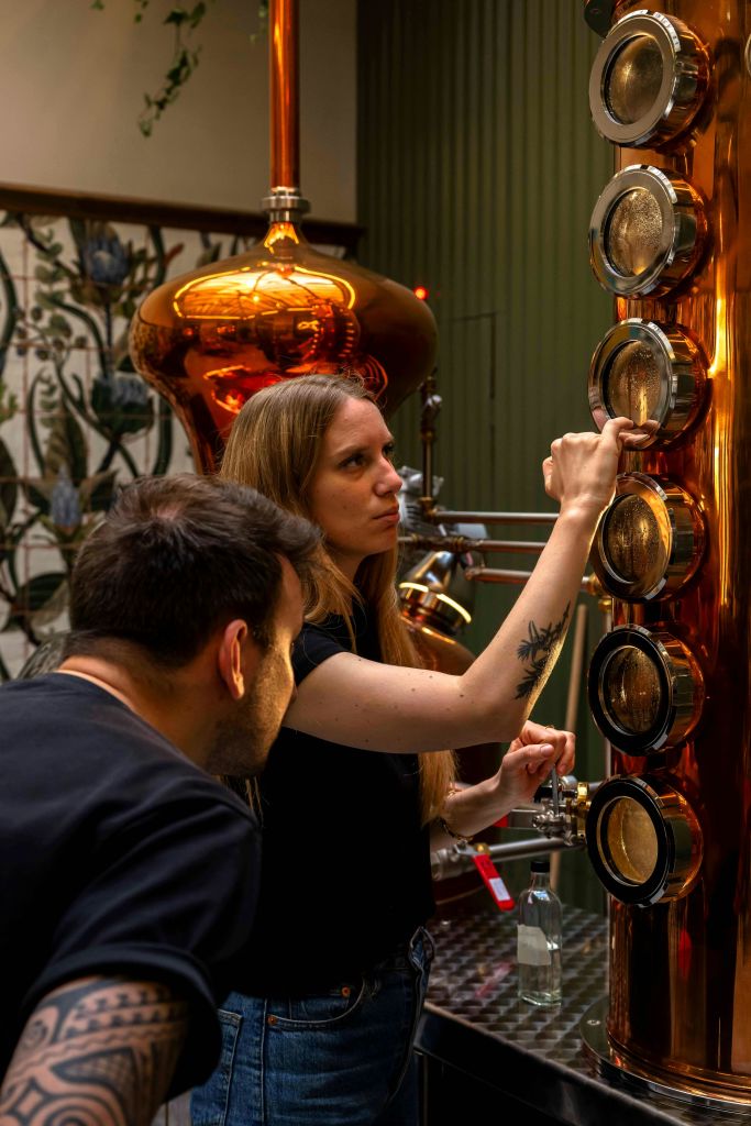 조명이 밝은 공간에 여러 개의 원형 보기 포트가 있는 구리 증류 장치를 검사하는 두 사람.