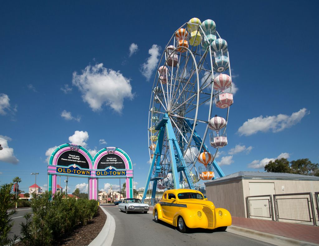 밝은 노란색 빈티지 자동차, 회색 자동차, "올드 타운" 입구 표지판이 있는 도로 위에 다채로운 대관람차가 있습니다.