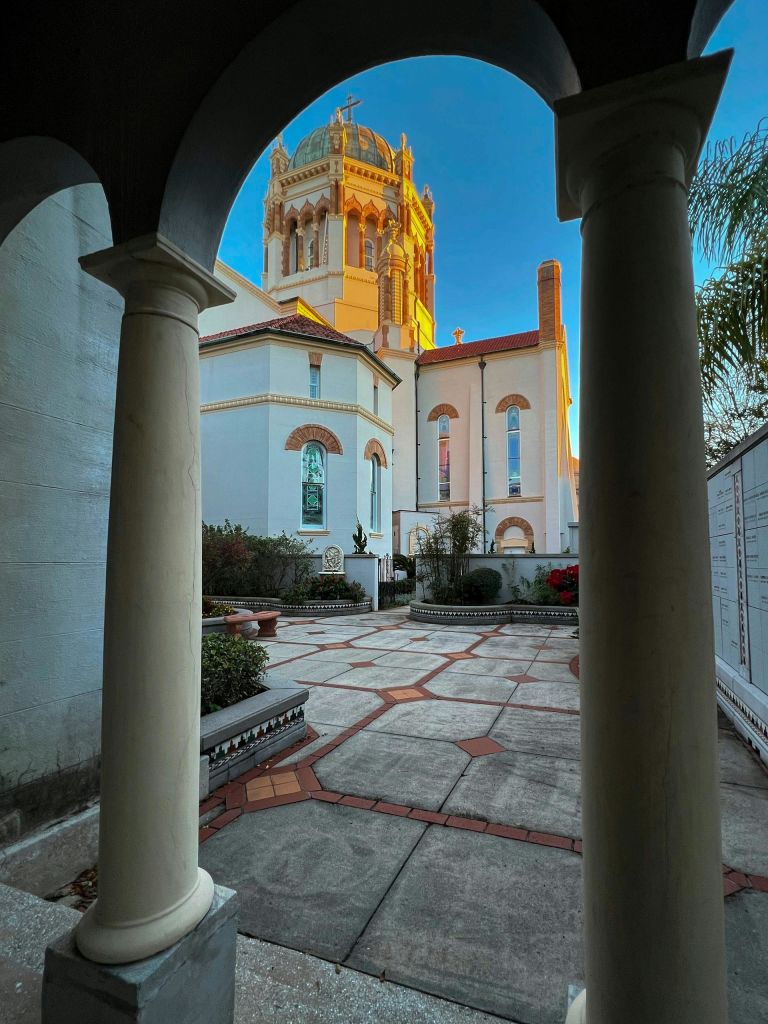 높은 흰색 기둥과 건물로 둘러싸인 타일 패턴의 안뜰로, 배경에 타워가 보입니다.