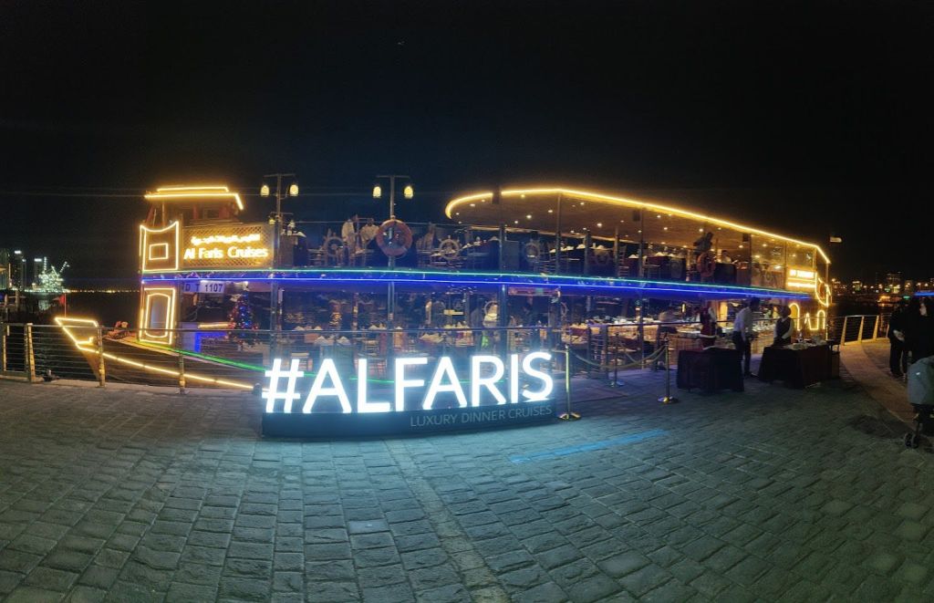 "ALFARIS Luxury Dinner Cruises" 표지판이 있는 밝은 조명의 보트는 밤에 장식과 사람들이 탑승한 채로 정박해 있습니다.