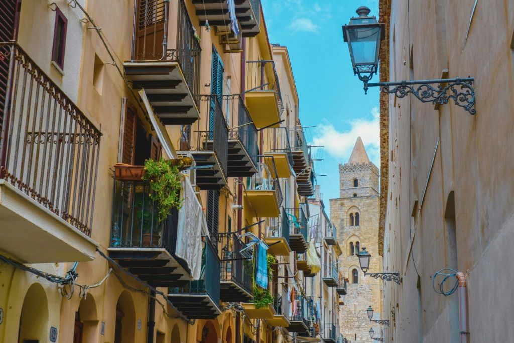 좁은 거리에 빨래가 달린 발코니가 있는 건물이 있습니다. 푸르고 부분적으로 흐린 하늘 아래 역사적인 탑이 보입니다.