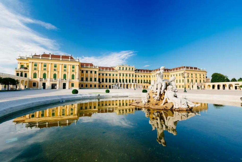 맑고 푸른 하늘 아래, 물에 비친 전경에 동상 분수가 있는 커다란 노란 궁전.