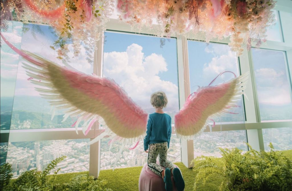 커다란 분홍색과 흰색 날개를 가진 어린이가 구름으로 가득한 하늘 아래 창밖의 도시 풍경을 바라보고 있습니다.