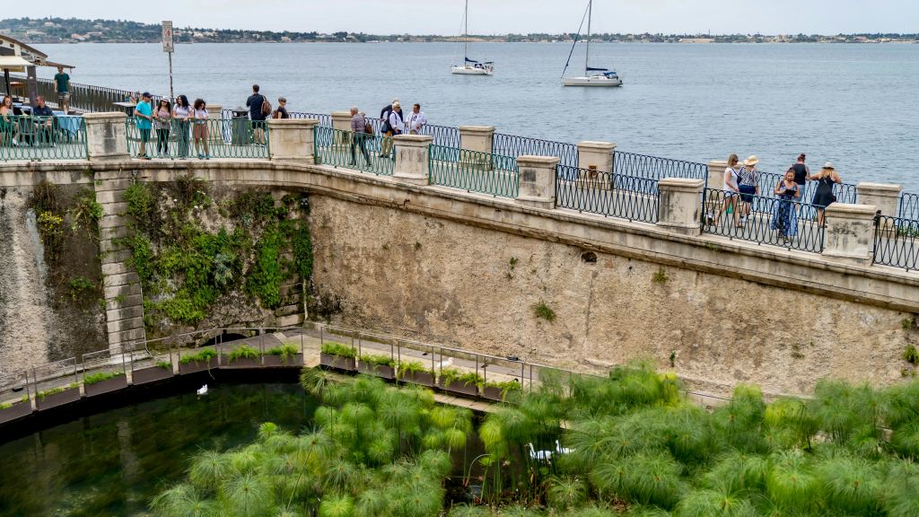 사람들은 요트와 함께 물줄기가 내려다보이는 석조 산책로를 따라 걷습니다. 앞쪽에는 푸른 식물이 연못을 둘러싸고 있습니다.