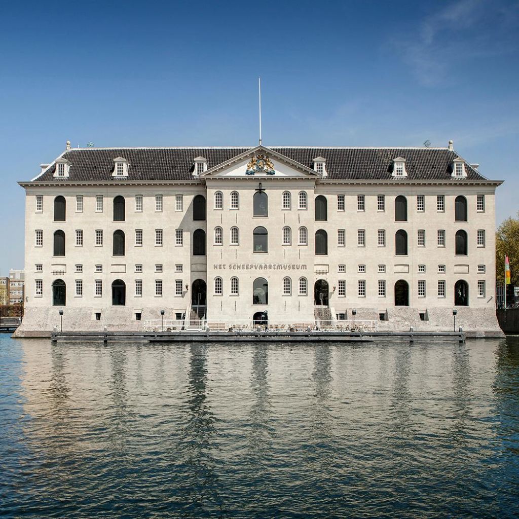 수많은 창문과 어두운 지붕이 있는 크고 유서 깊은 건물이 전경의 잔잔한 물에 반사되어 있습니다.