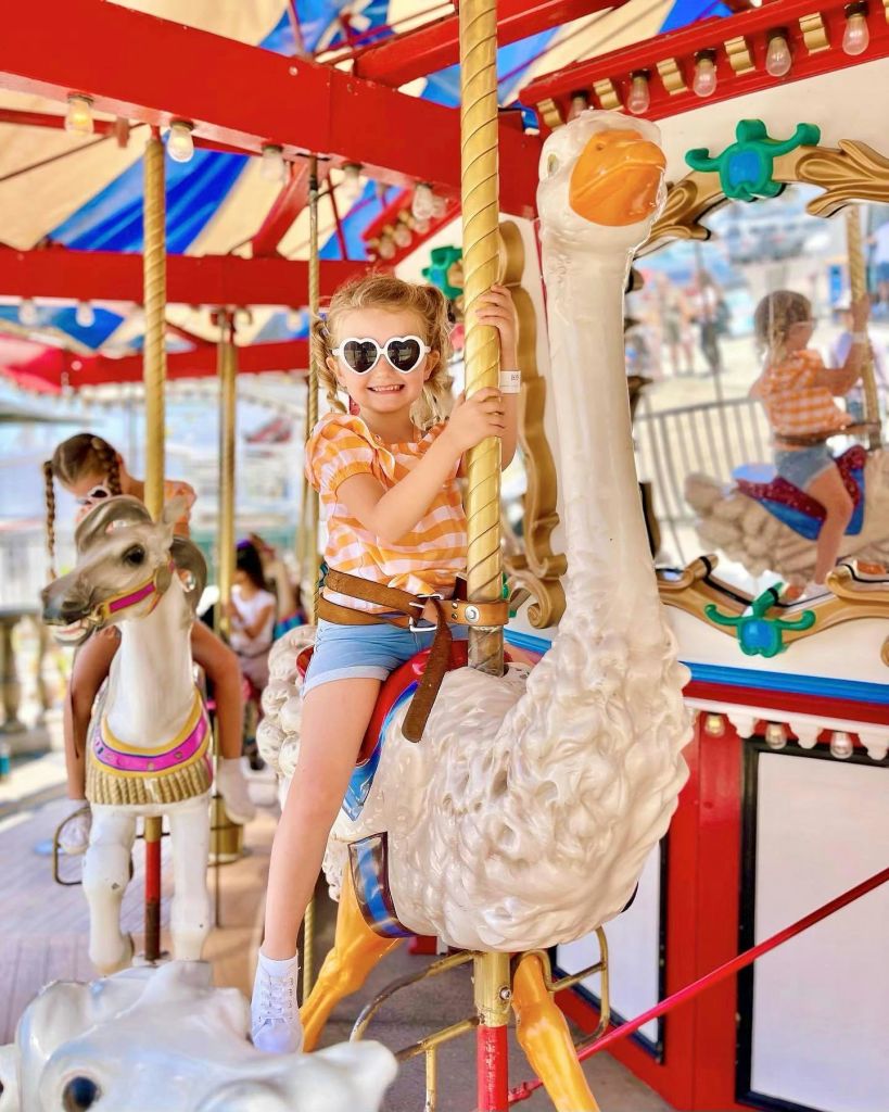 하트 모양의 선글라스를 쓴 어린 소녀가 회전목마를 타고 흰 새를 타고 환하게 웃고 있습니다. 어린이를 위한 차량 서비스가 늘어나고 있습니다.
