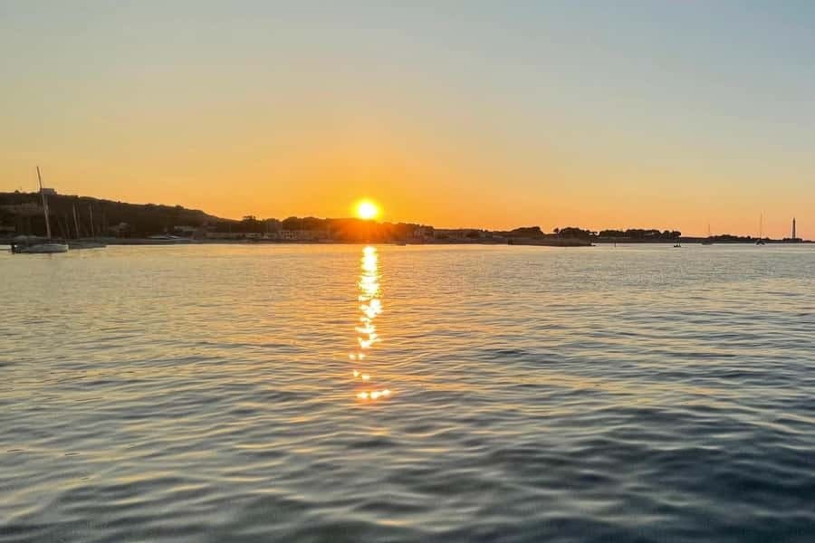 햇살이 반사되는 잔잔한 물 위에 지는 석양과 나무와 건물이 있는 먼 해안선.