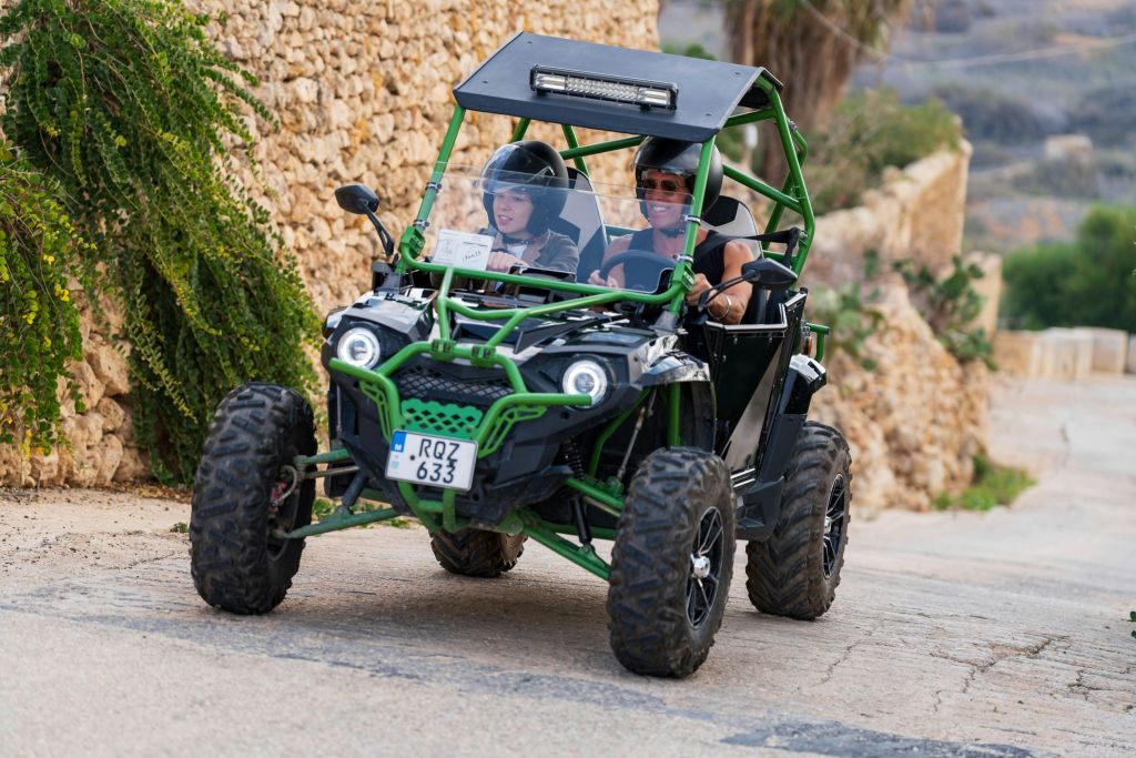 근처에 돌담과 식물이 있는 돌 포장 도로에서 녹색 오프로드 차량을 운전하는 두 사람이 헬멧을 쓰고 있습니다.