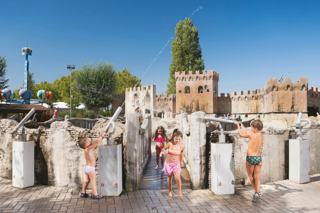 맑고 푸른 하늘 아래 성을 테마로 한 구조물과 워터젯이 있는 물놀이장에서 놀고 있는 어린이들.