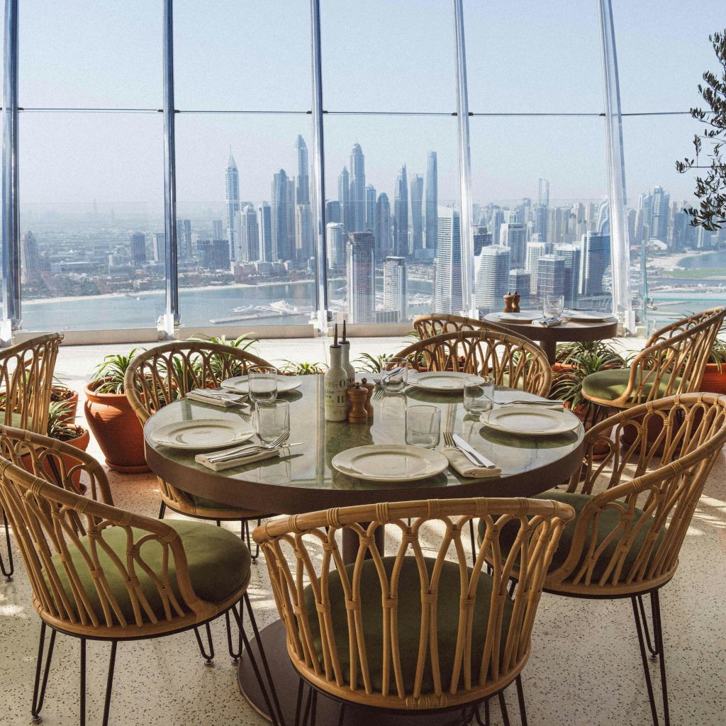 도시의 스카이라인이 내려다보이는 유리 벽으로 둘러싸인 레스토랑에 6개의 고리버들 의자가 놓인 원형 식탁이 있습니다.