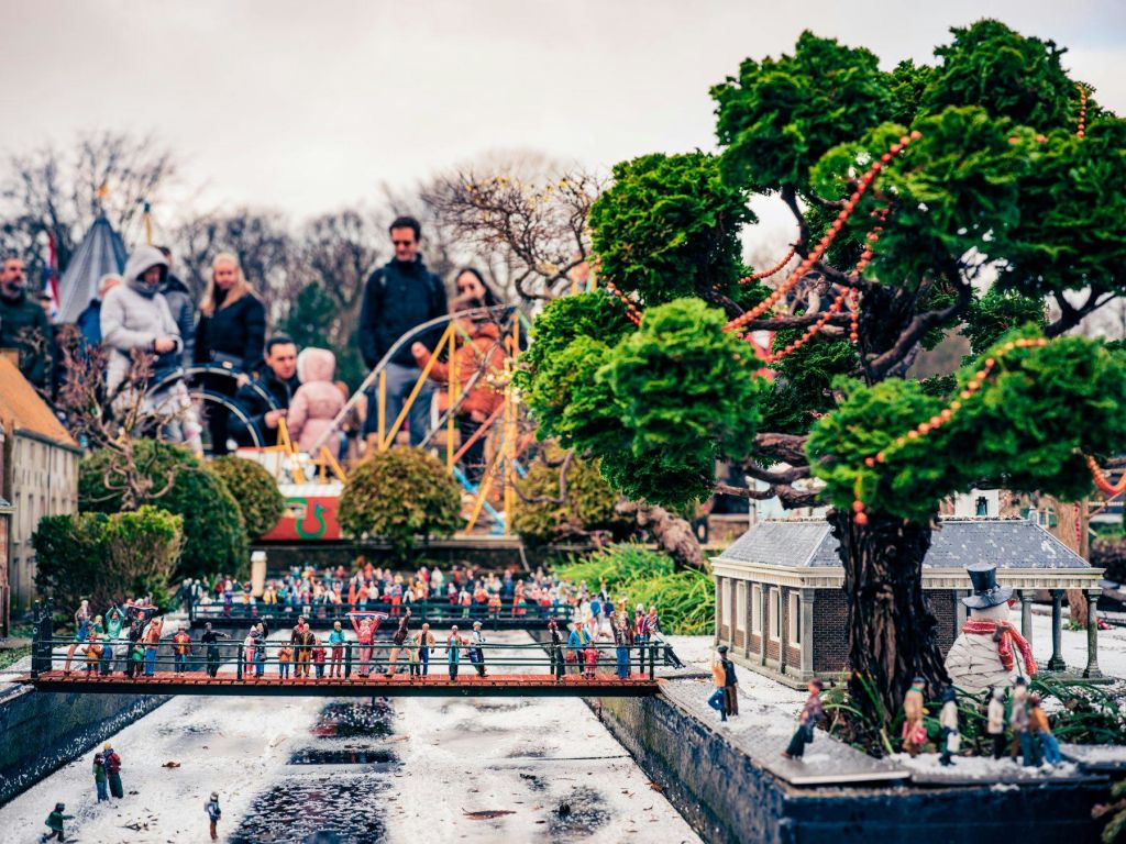 나무와 건물로 둘러싸인 운하를 가로지르는 다리에 모범생들이 앉아 있는 미니어처 장면, 배경이 흐릿한 실제 사람들.