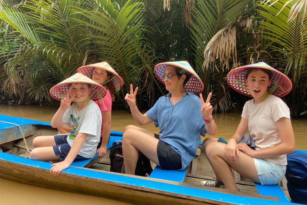 푸르른 단풍을 배경으로 활짝 핀 모자를 쓴 세 사람이 강에서 보트를 타고 있습니다.