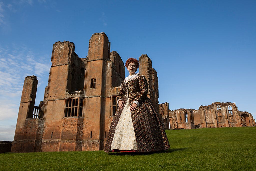 맑은 하늘 아래 잔디밭에 있는 낡고 부분적으로 폐허가 된 벽돌 건물 앞에 서 있는 마침표 의상을 입은 사람.
