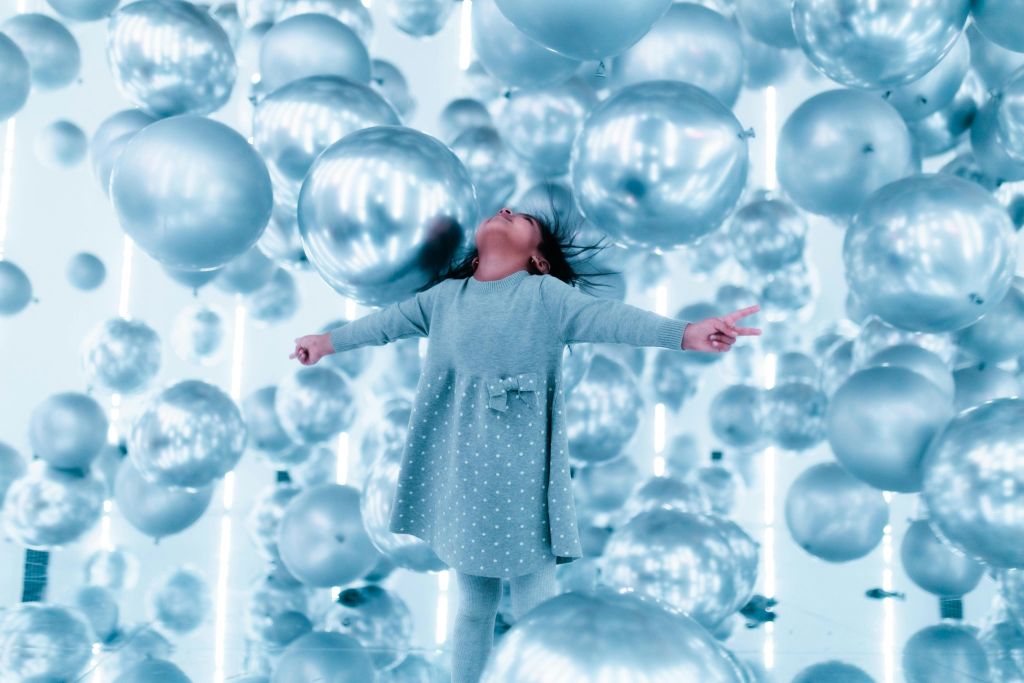 회색 옷을 입은 어린이가 밝고 반사되는 공간에서 수많은 은색 풍선 사이에서 즐거운 포즈를 취하고 있습니다.