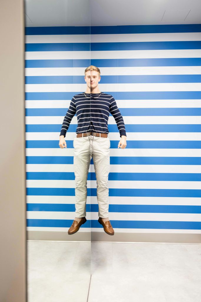 한 사람이 가로 파란색과 흰색 줄무늬가 있는 벽 앞에 서서 줄무늬 셔츠와 흰색 바지를 입고 있습니다.