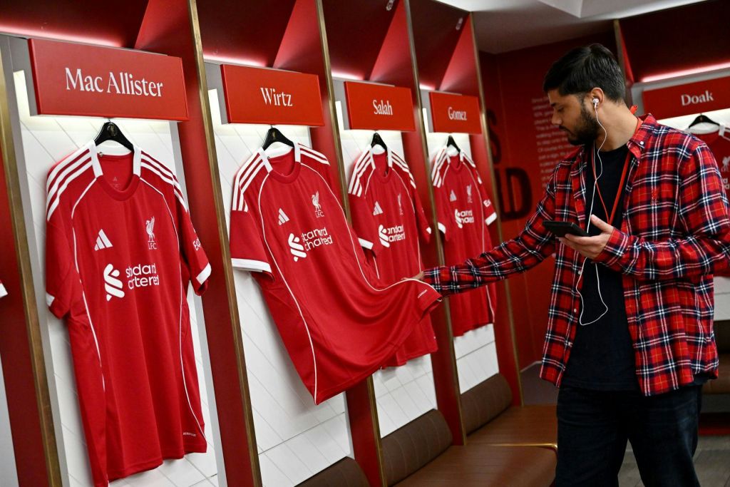 빨간색 격자무늬 셔츠를 입은 한 남성이 맥 앨리스터, 위르츠, 살라, 고메즈라는 라벨이 붙은 다른 세 개의 매달린 유니폼이 있는 라커룸에서 빨간색 리버풀 FC 유니폼을 검사하고 있습니다.