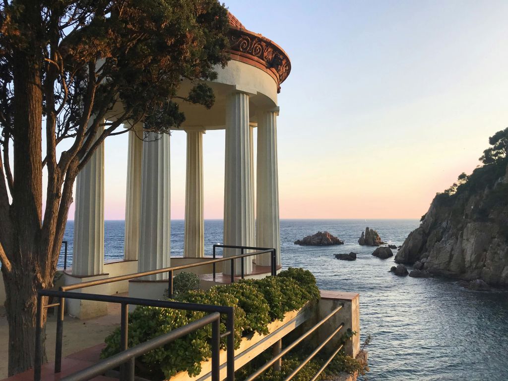 석양이 질 무렵 바다가 내려다보이는 기둥형 파빌리온으로, 앞쪽에는 바위 노두와 녹음이 우거져 있습니다.